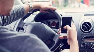 A person drives a car while holding a smartphone, suggesting distracted driving. The focus is on the hand with the phone, highlighting a safety concern.