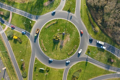 Aerial view of a green roundabout with cars navigating the circular roadway. Bright sunlight casts shadows, and grassy areas are neatly manicured.
