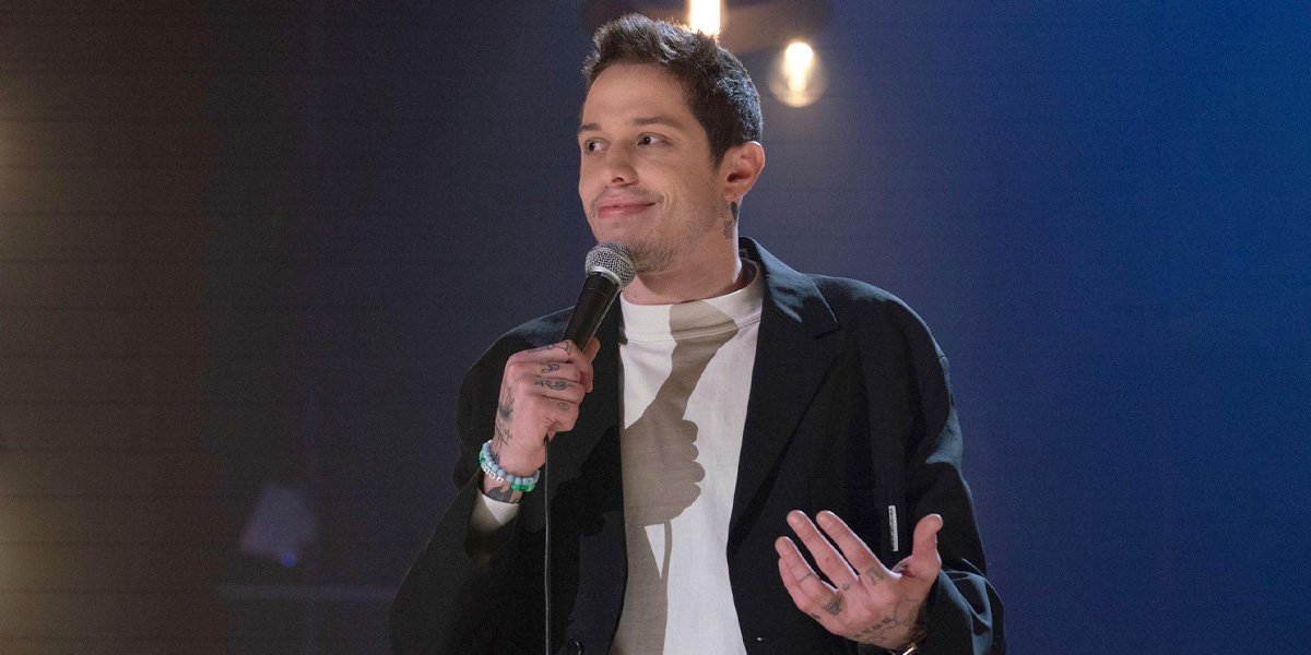 A man with short brown hair holds a microphone and gestures while performing stand-up comedy on stage, wearing a black jacket over a light shirt, with stage lights in the background.
