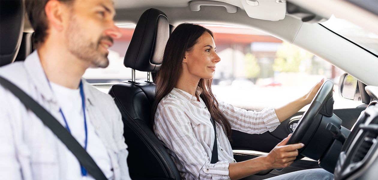 A woman is driving a car while a man sits in the passenger seat beside her. Both are wearing seat belts, and sunlight is shining through the windows. The woman looks focused on the road ahead.