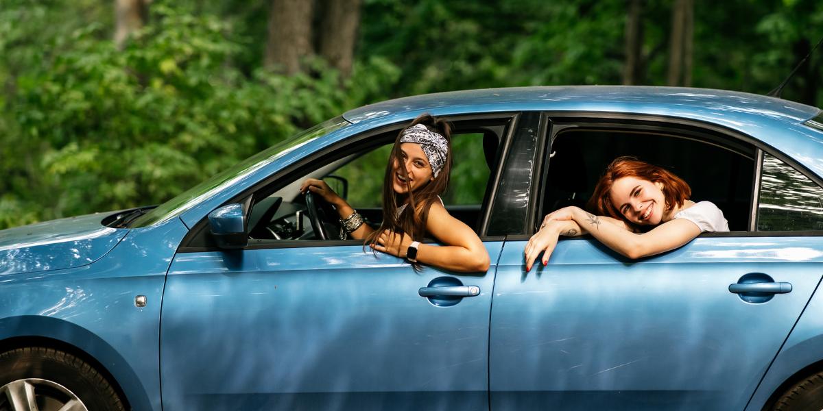 Two smiling women in a blue car enjoy a forest drive. One leans out the driver’s window, the other rests on the backseat window, exuding a carefree vibe.